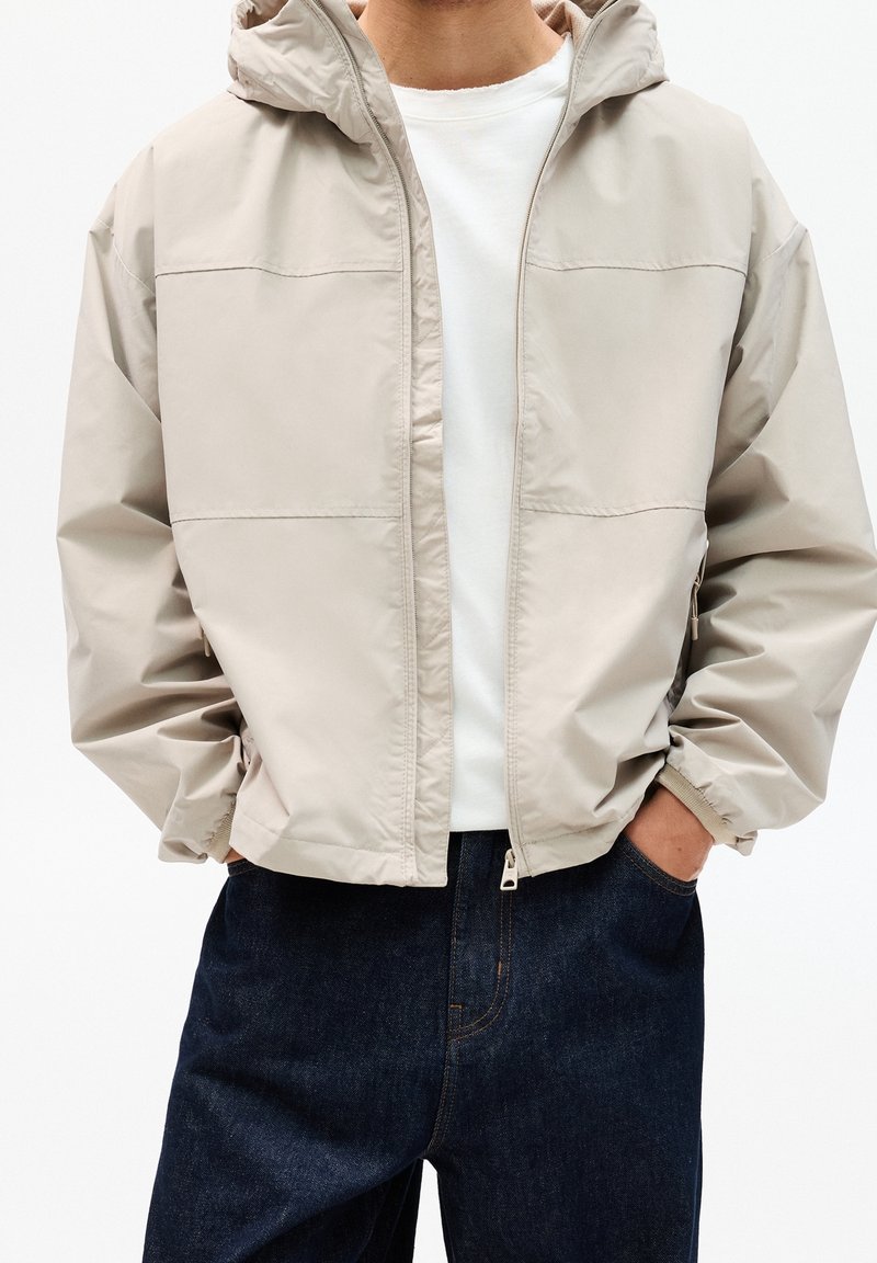 Personne portant une veste à capuche beige zippée par-dessus une chemise blanche, les mains dans les poches d’un jean en denim bleu foncé.