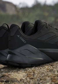 Zwarte Terrex trailrunningschoenen met robuuste zolen, geplaatst op rotsachtig terrein buiten.