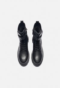 Bottines en cuir noir avec un design à lacets, doublure synthétique, semelles épaisses et sangles décoratives au niveau de la cheville. Texture de surface lisse.