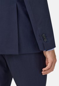 Mornarsko moder suknjič z teksturiranim zaključkom, ki prikazuje razporek in štiri črne gumbe na manšetah, nosi pa se ga čez svetlo moder karirasti srajc.