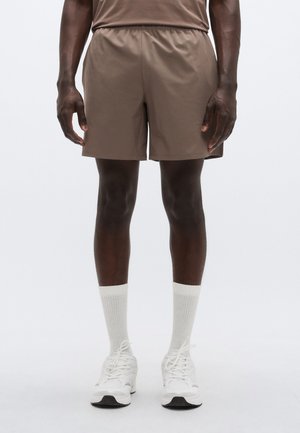 Pruunid spordipüksid elastse vöökoha, sileda tekstuuri ja põlvepikkuse lõikega, kantud valgete sokkide ja tossudega.