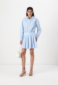 Vestido de camisa riscado azul claro com mangas longas, cintura franzida e saia plissada. Combinado com sandálias de salto bege e uma pequena mala branca.