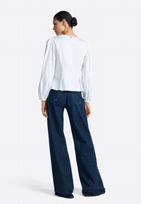 Balta blūze ar pumpainām piedurknēm, piegulošu vidukli un aizmugurējo pogu detaļu, kopā ar tumši zilām, plašām džinsu biksēm ar standarta šuvēm.