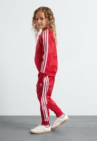 Chándal deportivo rojo con rayas blancas, que incluye una chaqueta con cremallera y pantalones ajustados. Combinado con zapatillas blancas con suelas de goma.