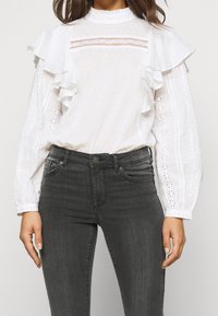 Weiße Bluse mit Rüschenärmeln, strukturiertem Stoff und Spitzenakzenten; kombiniert mit dunkelgrauen, enganliegenden Jeans. Ausführliche Lochmuster entlang der Ärmel.