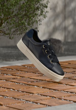 Zapatilla deportiva de cuero negro con suela blanca y cordones negros, flotando sobre un suelo de baldosas de madera al aire libre con un fondo desenfocado de árboles y una pared.