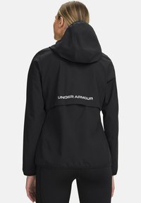 Giacca impermeabile nera con cappuccio, caratterizzata da un logo bianco "UNDER ARMOUR" sul retro e un design ampio e non strutturato.