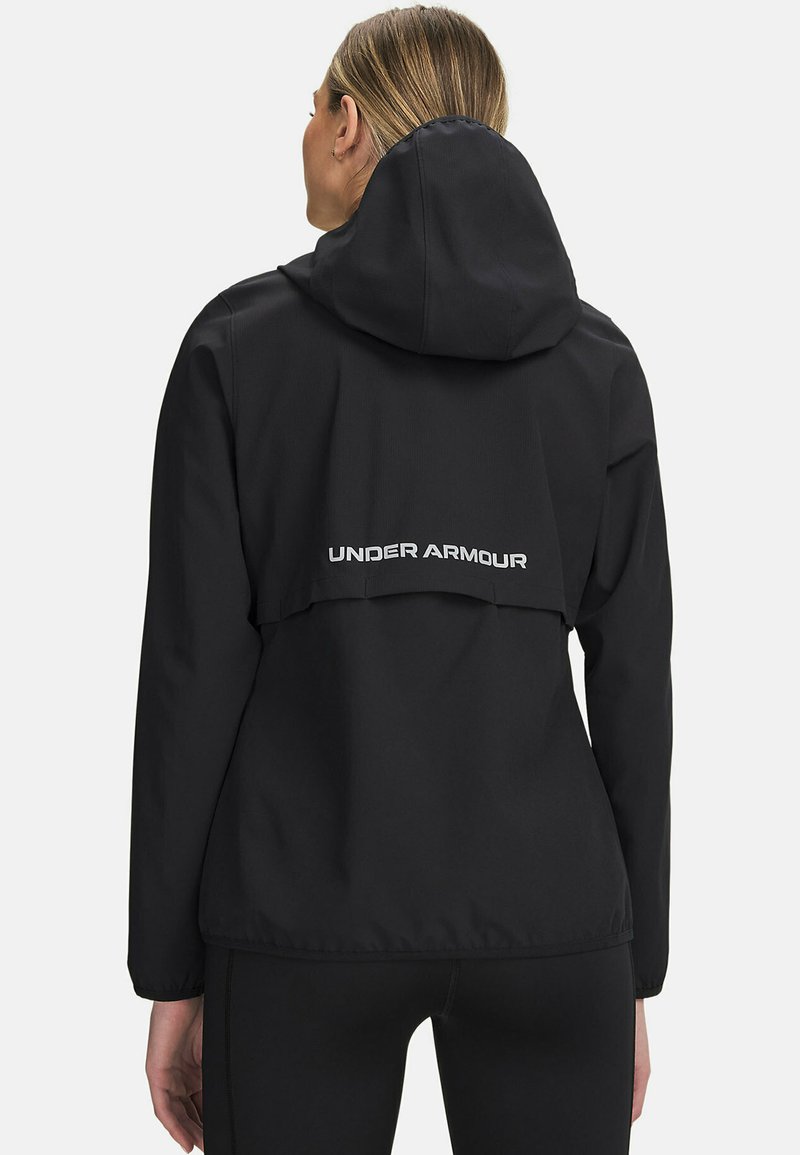 Giacca impermeabile nera con cappuccio, caratterizzata da un logo bianco "UNDER ARMOUR" sul retro e un design ampio e non strutturato.