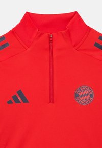 Κόκκινο αθλητικό πάνω μέρος με φερμουάρ στο ένα τέταρτο, υψηλός γιακάς, λογότυπο FC Bayern München και οι τρεις ρίγες της Adidas σε σκούρο γκρι. Λεία υφή υφάσματος.