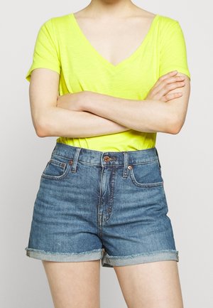 Personne portant un t-shirt jaune vif à col en V et un short en jean bleu roulé, les bras croisés sur la poitrine.