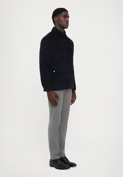 Marineblaue Cordjacke mit zwei Brusttaschen und einem Reißverschluss, kombiniert mit hellgrauen Hosen und schwarzen Desserschuhen.