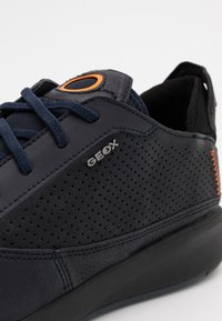 Baskets bleu marine avec des panneaux latéraux perforés, design à lacets, logo Geox en argent et accent orange sur la languette, avec un matériau texturé.