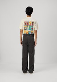 Camiseta de color beige claro con un diseño gráfico colorido en la parte posterior, combinada con pantalones de pierna ancha gris oscuro que presentan un acabado texturizado.