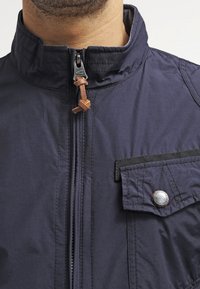 Navyblauwe lichte jas met een hoge kraag, ritsluiting, leren accent en een voorklepzak met een drukknop.