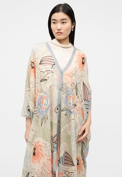 Γυναίκα με μακριά μαύρα μαλλιά φοράει ένα ανοιχτόχρωμο, με κουμπιά, διάφανο φόρεμα με λουλουδένιο μοτίβο πάνω από ένα εκρού ζιβάγκο, σε λευκό απλό φόντο.