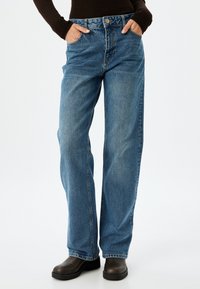 Jeans de mezclilla azul claro, de pierna ancha y cintura alta, con dos bolsillos delanteros y cierre de botón. Llevados con calzado oscuro.