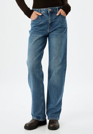 Helle blaue, weite Jeans aus Denim mit hoher Taille, zwei frontalen Taschen und einem Knopfverschluss. Getragen mit dunklem Schuhwerk.