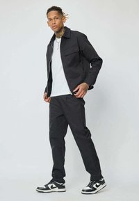 Homme debout portant une veste noire, une chemise blanche, un pantalon noir et des baskets noir et blanc devant un fond uni.