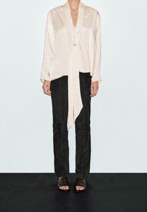 Femme portant un chemisier ample de couleur crème avec un long ruban, un pantalon droit noir et des sandales noires à bouts ouverts, debout devant un fond uni.