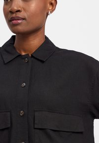 Zwarte button-up overhemd met korte kraag, voorzien van twee borstzakken en bruine knopen. De stof lijkt glad met een lichte glans.