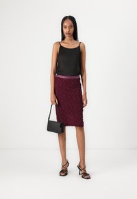 Schwarzes Satin-Camisole mit dünnen Trägern, kombiniert mit einem bordeauxfarbenen, strukturierten Bleistiftrock und einer schwarzen Handtasche. Das Modell trägt schwarze High Heels mit Riemchen.