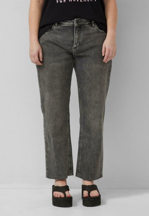 Osoba ubrana w wyblakłe czarne proste dżinsy z postrzępionymi nogawkami i czarne sandały na platformie, stojąca na tle jednolitej szarej ściany.