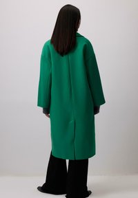 Roheline villane segu jakk laia silueti, laiate varrukate ja taga lõhiku detailiga. Kandmine tumedate laiapõhjaliste pükste ja tekstuuriga halli aluspuseriga.