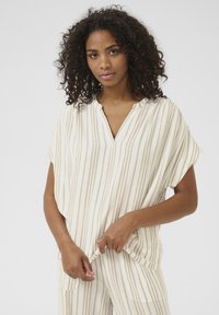 Blouse légère en crème avec des rayures verticales beiges, manches courtes et un décolleté en V. Texture douce, coupe décontractée et minimalisme dans les détails.