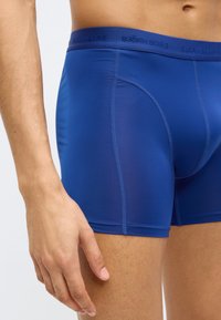 Μπλε μποξέρ σλιπ από λείο, ελαστικό υλικό. Διαθέτουν μονόχρωμο σχέδιο, ζώνη μέσης με λογότυπο και επίπεδες ραφές για άνετη και αεροδυναμική εφαρμογή.