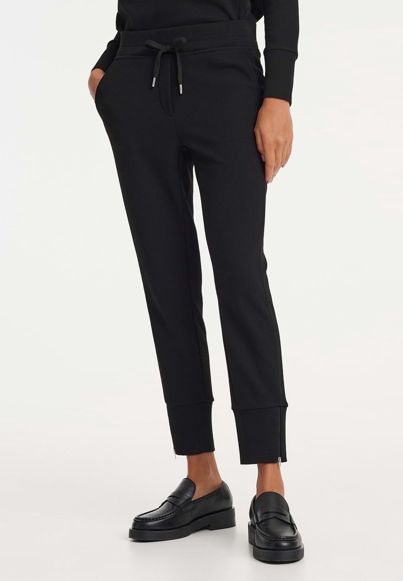 Personne portant un pantalon de jogging noir avec poches avant, taille à cordon, fermetures éclair aux chevilles, et des mocassins noirs sans lacets, debout devant un fond blanc.
