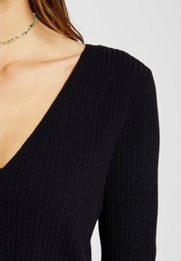 Top largo de manga larga negro con un diseño de escote en V profundo, que destaca por su textura suave y su silueta ajustada. Collar tipo choker con cuentas multicolores.
