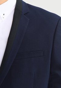 Mörkblå kostymjacka med svart slag som bärs över en vit skjorta med knäppt krage.