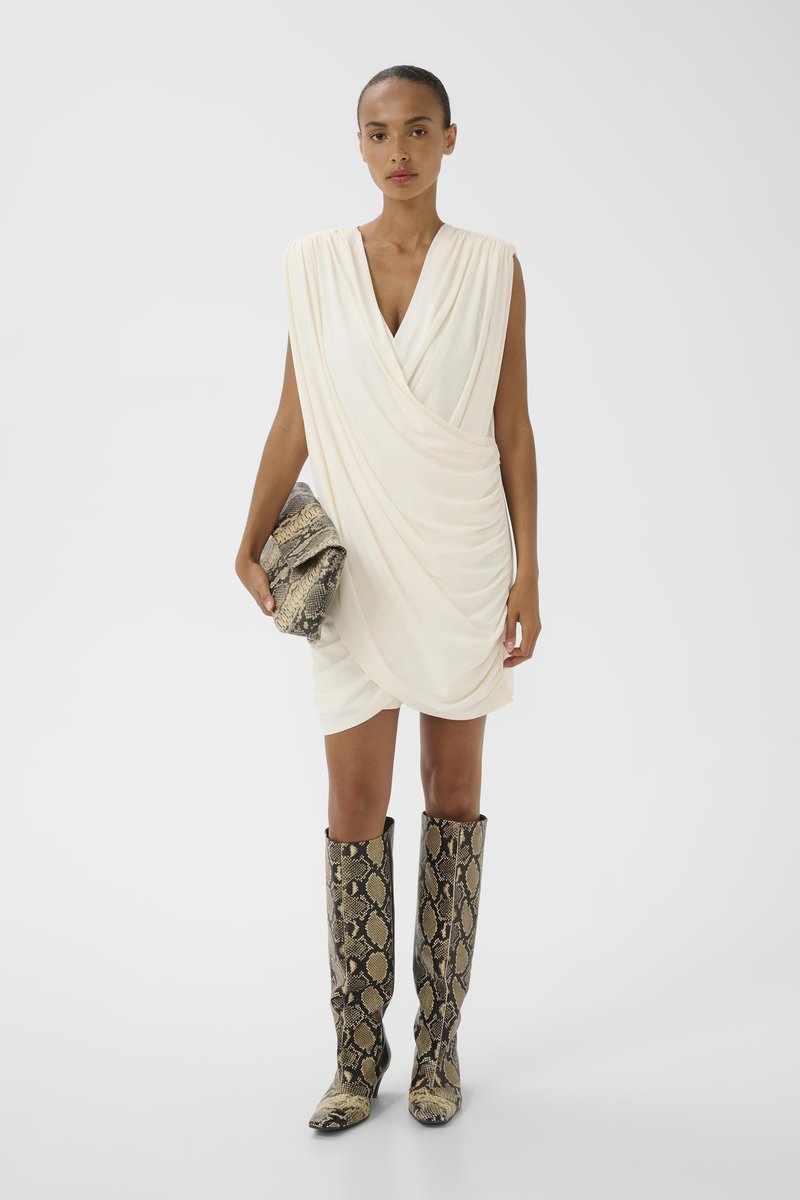 Femme portant une mini-robe blanche sans manches drapée, tenant une pochette en peau de serpent et portant des bottes hautes assorties en peau de serpent.