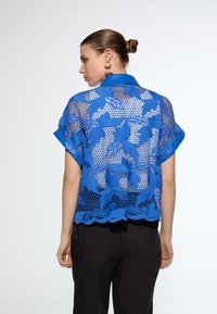 Blue lace shirt with floral pattern, short sleeves, and a collared neckline. Lightweight and semi-transparent, featuring scalloped hemline.