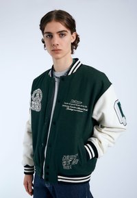 Giacca varsity verde e crema con dettagli ricamati, bottoni a scatto e polsini a righe. Presenta testo e un grande grafico sul davanti.