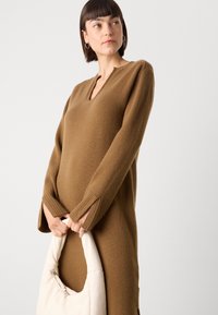 Abito a maglia marrone con scollatura a V e polsini a costine. Il modello tiene una borsa a spalla bianca. Tessuto texturizzato, design a lunghezza media.