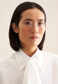 Blusa bianca con un ampio colletto e un dettaglio al collo annodato. Tessuto dalla texture liscia, design aderente, viso parzialmente visibile, sfondo neutro.