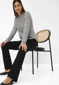 Grijze gebreide trui met sprankelende details, zwarte op maat gemaakte broek en platte zwarte schoenen, zittend op een zwarte stoel met een geweven rotan rugleuning.