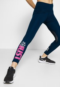 Laivameren siniset leggingsit, joissa on vaaleanpunainen "JUST"-teksti ja hopea logo. Korkeavyötäröiset, joustavaa materiaalia, siro muotoilu ja muotoilevat saumat.