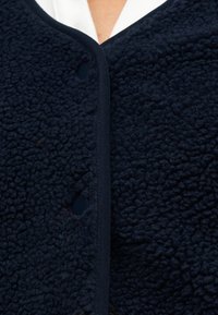 Κοντινό πλάνο μπροστινού μέρους σκούρου ναυτικού μπλε υφασμάτινης ζακέτας με υφή fleece και κουμπιά, φορεμένη πάνω από λευκό πουκάμισο.