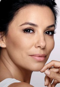 Gros plan sur un visage avec une peau lisse de couleur marron clair. Les traits incluent des cils foncés et fournis, une couleur de lèvres naturelle et un léger fard aux joues. Bijoux simples visibles.