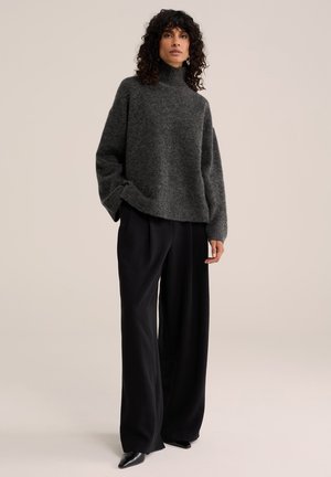 Femme aux cheveux bouclés et foncés portant un pull gris à col roulé ample, un pantalon noir large et des chaussures noires pointues, debout devant un fond uni.