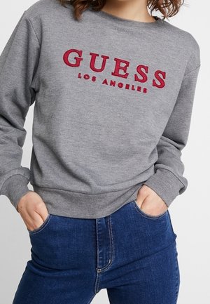Osoba ubrana w szarą bluzę z czerwonym napisem „"GUESS LOS ANGELES"” i niebieskie dżinsy, z rękami w kieszeniach, stoi na jednolitym tle.
