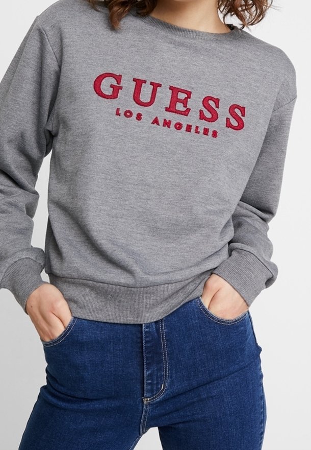 Personne portant un sweat gris avec le texte rouge « "GUESS LOS ANGELES" » et un jean bleu, les mains dans les poches, debout devant un fond uni.