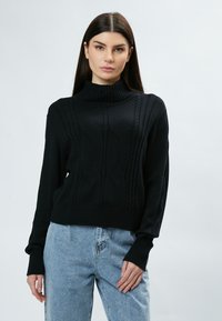 Schwarzer gestrickter Rollkragenpullover mit Zopfmuster, gerippten Bündchen und cropped Design, kombiniert mit hoch taillierten blauen Jeans.