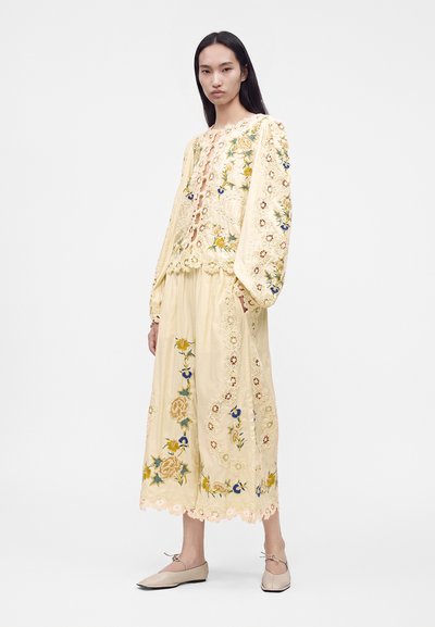 Woman wearing a pale yellow embroidered blouse and skirt set with floral patterns and scalloped lace edges, paired with beige flats.