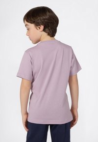 Niño con cabello corto marrón, vestido con una camiseta corta de color lila claro y pantalones oscuros, girado ligeramente hacia un lado contra un fondo liso.