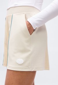 Κοντινό πλάνο ενός ατόμου που φοράει μπεζ σορτς με το χέρι στην τσέπη, μια κάθετη μπλε ρίγα και ένα λευκό οβάλ λογότυπο κοντά στο τελείωμα.