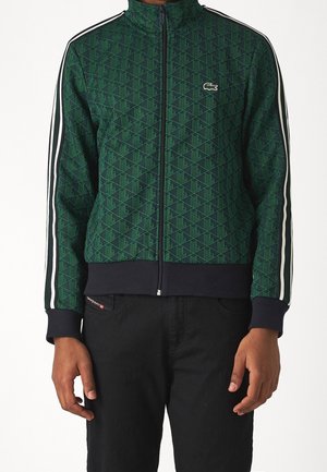 Hombre con chaqueta verde con estampado y cremallera, con franjas laterales blancas, y pantalones negros, sobre un fondo blanco.