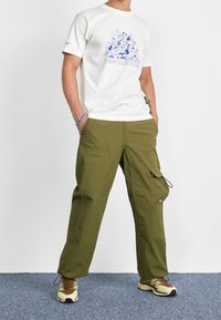 Man i vit grafisk New Balance t-shirt, olivgröna cargobyxor och beige bruna löparskor står på en blå matta.